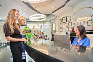 A woman and a child at a clinic reception, with the child holding a green toy and the woman smiling while standing in front of a reception desk with a laptop and a flower vase.