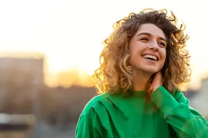 A woman with curly blonde hair is smiling while looking forward with a bright light on her face