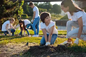 Five children and one adult female are planting trees in a grassy area. One child is kneeling and holding a shovel, and the others are watching.