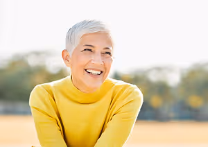 A woman with white hair is smiling and posing for a photo outside in a field with trees in the background.