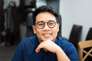 A smiling man with glasses sits in a chair with his chin resting on his hand.