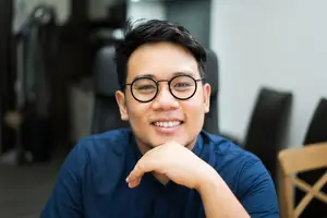 A smiling man with glasses sits in a chair with his chin resting on his hand.