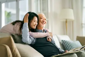 An elderly man and a young woman are sitting on a couch and smiling while reading a book together.