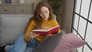 A woman is sitting on a couch and reading a book while wearing glasses and a yellow sweater