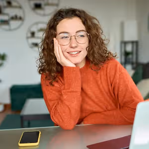 A woman is sitting at a desk, smiling, with a phone and laptop in front of her.