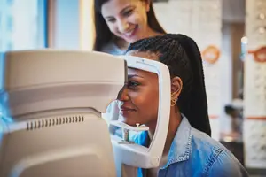A woman is looking into a white eye exam machine while another woman smiles and watches her in a room with a window and white walls