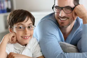A young boy wearing glasses and a blue shirt is sitting next to a smiling man who is also wearing glasses and a blue shirt.