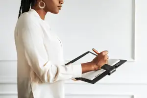Woman writing on a notepad with a pen while standing in front of a white wall