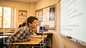 a young boy with glasses is sitting in a classroom with other students, a whiteboard and bulletin boards in the background, with a text overlay that reads Vision First Eye Care