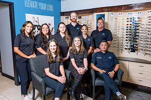 A group of people in black polo shirts and pants, standing in a room with a blue wall and a blue board with the words Elevate Your Look. Some are sitting on chairs while others are standing behind them. There is a glass display with sunglasses and a wooden cabinet with drawers in the room.