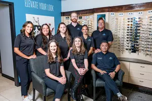 A group of people in black polo shirts and pants, standing in a room with a blue wall and a blue board with the words Elevate Your Look. Some are sitting on chairs while others are standing behind them. There is a glass display with sunglasses and a wooden cabinet with drawers in the room.