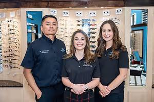 Three people are smiling and posing for a photo in front of an eyeglass store.