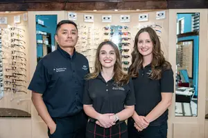 Three people are smiling and posing for a photo in front of an eyeglass store.