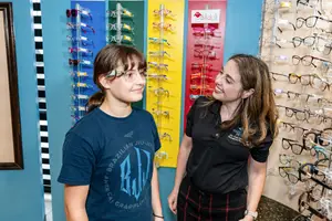 Two women standing in front of a display of eyeglasses, one of them wearing glasses and smiling while the other woman is looking at the glasses on the wall.