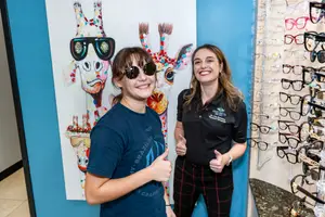 Two women are standing in front of a colorful giraffe poster and a rack of glasses, smiling and posing for a photo.