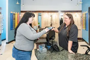 Two women are standing behind a reception desk in an optical shop, one of them is holding a device and they are both smiling