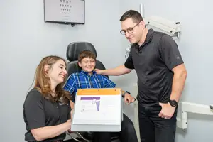 A young boy is being held by a woman and a man who are both smiling, the boy is holding a folder with some text and an image on it