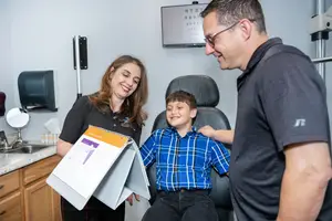 A family of three is in an optometrist's office, the man and woman are smiling while the boy is looking at the camera, the woman is holding a folder with some papers inside.