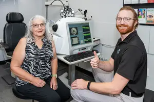 A woman and a man are sitting in front of an eye examination machine, both smiling and posing for a photo.