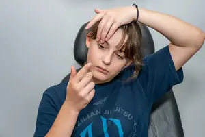 A young person wearing a Brazilian Jiu Jitsu t-shirt and a bracelet removes a contact lens from their eye.