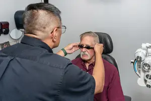 An optician is examining an older man wearing glasses and a maroon shirt in a room with a white wall and medical equipment.