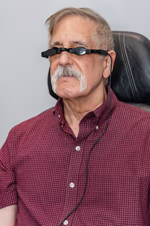 A man with a white mustache and beard is sitting in a chair with a black leather seat. He is wearing a maroon checkered shirt and has a black cord attached to his eyeglasses. He is looking to the left, and his eyes are closed. The background is white, and he is in a room.