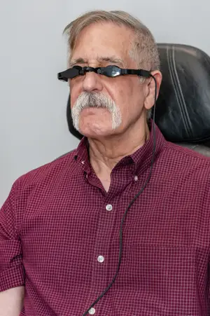 A man with a white mustache and beard is sitting in a chair with a black leather seat. He is wearing a maroon checkered shirt and has a black cord attached to his eyeglasses. He is looking to the left, and his eyes are closed. The background is white, and he is in a room.