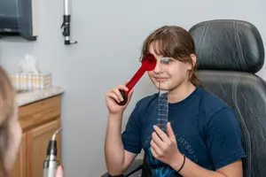 A girl with glasses is holding a red magnifying glass on her face while a person is standing in front of her holding a black object