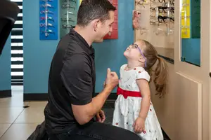 A man in a black shirt is kneeling next to a young girl wearing glasses in an optician's shop.