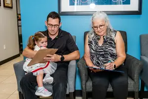 A man, woman, and young girl are sitting in a waiting room. The man and woman are holding clipboards, and the man is looking at the young girl.