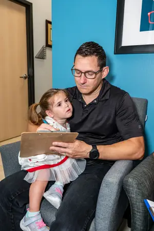 A man wearing glasses and a watch is sitting on a couch with a little girl. They are both holding a clipboard and looking at something on it. The man is wearing a polo shirt and the little girl is wearing a dress with a red ribbon on her ponytail. There is a picture frame on the wall behind them.