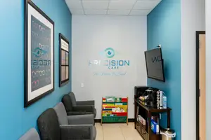 A Precision Care waiting room with blue walls, gray chairs, a coffee machine, a bookshelf, and a mounted monitor.