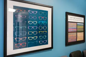 Two framed posters hang on a blue wall, one showing multiple pairs of colorful glasses and the other with a message about binocular vision disorder