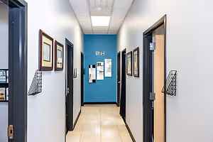 A hallway with white walls and blue accents, featuring framed pictures, a tiled floor, and an open door with an exit sign and a mirror.