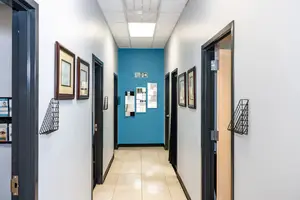 A hallway with white walls and blue accents, featuring framed pictures, a tiled floor, and an open door with an exit sign and a mirror.