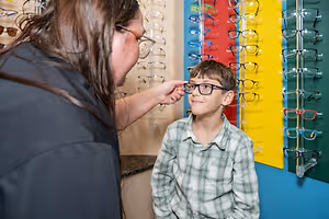 A woman is adjusting the glasses of a boy standing in front of a wall with glasses on display
