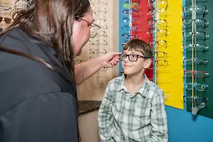 A woman is adjusting the glasses of a boy standing in front of a wall with glasses on display