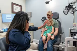 A man and a child are sitting in a chair in an eye clinic with a woman holding a camera.