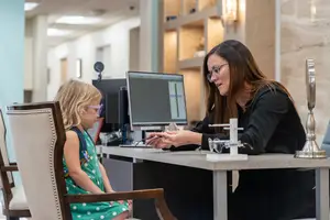 A woman with glasses is examining a young girl's eyes with a tool in a clinic setting.