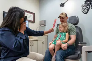 In an examination room, a man is sitting with a young girl while a woman is holding a device and checking the girl's eyes.