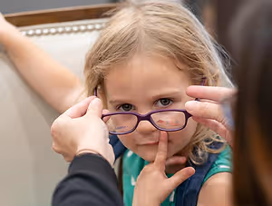 a little girl with blonde hair wearing purple glasses is looking at someone who is adjusting her glasses