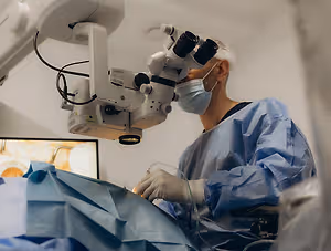 An eye doctor is performing a surgery with an ophthalmoscope while wearing a surgical mask and blue gown.