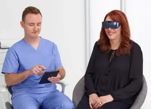 A man in a blue scrub is sitting in a chair and looking at a tablet while a woman in a black blouse is sitting next to him and wearing glasses.