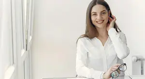 A smiling woman with brown hair wearing a white long-sleeve shirt and holding a pair of glasses in her hand