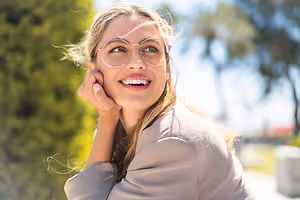 A woman with blonde hair and glasses is smiling and posing for a photo in a park with green bushes and trees