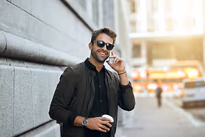 A man wearing sunglasses and a jacket is standing on the sidewalk and talking on his phone.