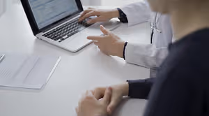 A person in a white lab coat with a stethoscope around their neck types on a laptop while another person looks on.