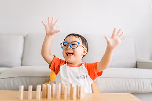 A child wearing glasses is playing with wooden blocks and has his hands raised in the air in a living room