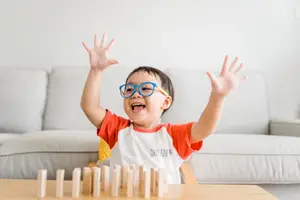 A child wearing glasses is playing with wooden blocks and has his hands raised in the air in a living room