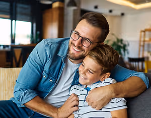 A man and a boy are sitting on a couch, smiling and looking at each other.